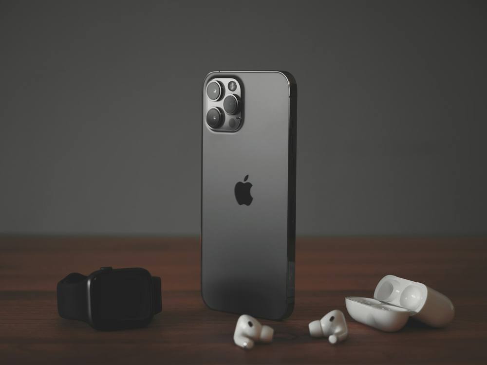 iPhone em pé sobre uma mesa de madeira, entre um Apple Watch e um par de AirPods